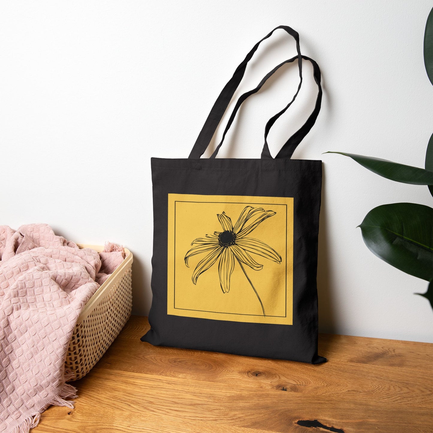 Black tote bag with a yellow flower design on a wooden surface with a white wall background