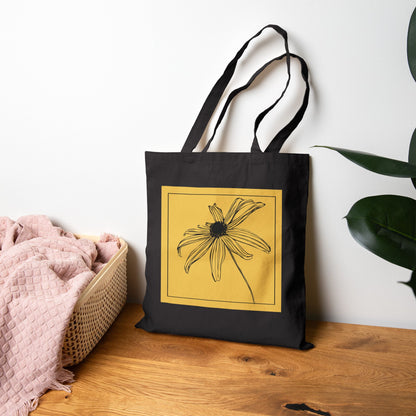 Black tote bag with a yellow flower design on a wooden surface with a white wall background