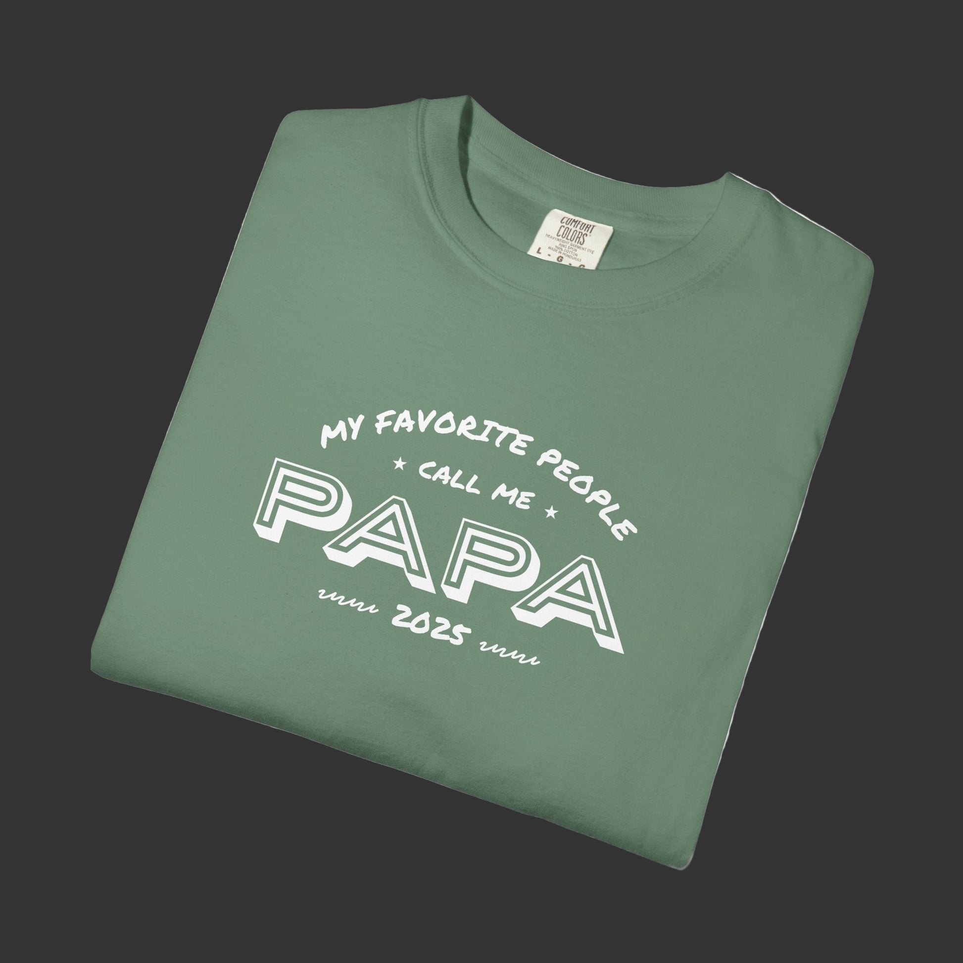 Folded green t-shirt with 'My Favorite People Call Me Papa 2025' text on a white background