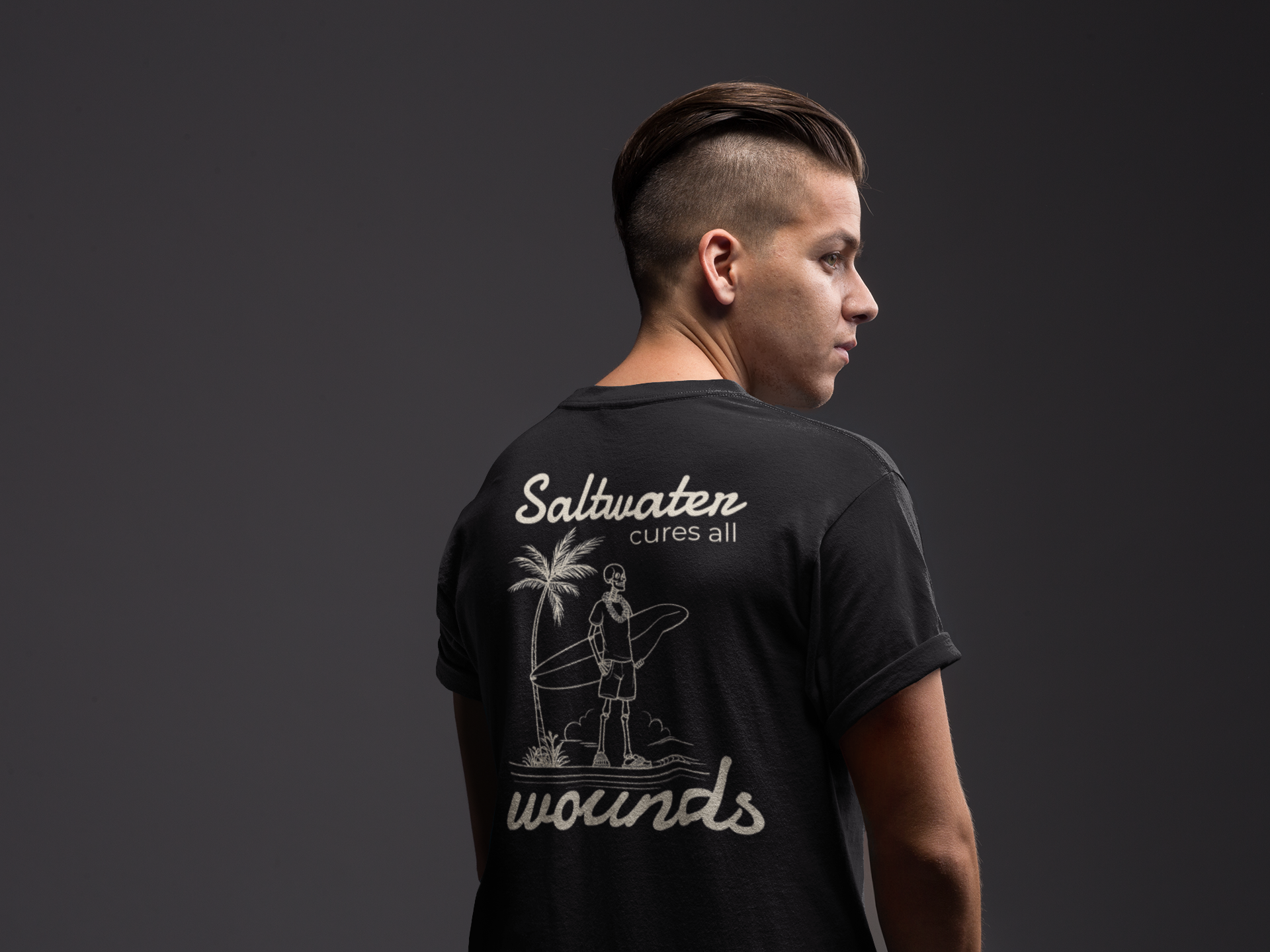 Person wearing a black t-shirt with 'Saltwater cures all wounds' text and graphic on the back against a dark background